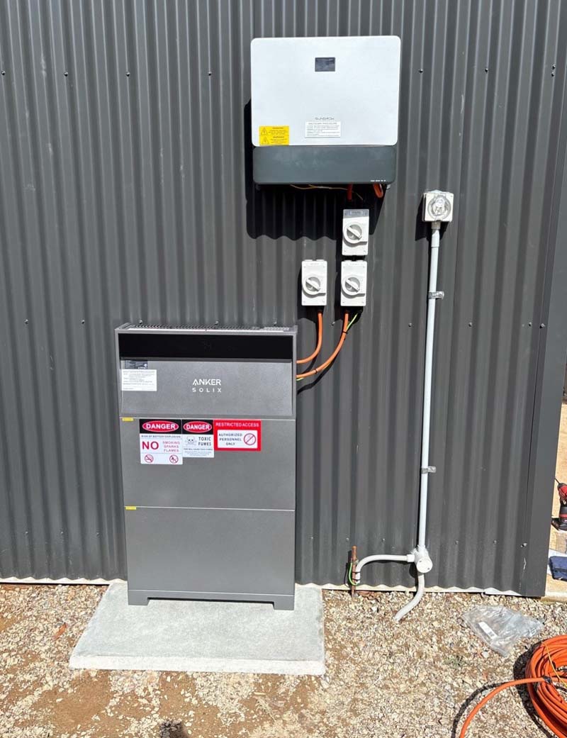 Outdoor electrical panel and inverter mounted on a corrugated metal wall, with wired conduits and warning labels.