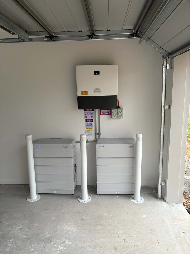 Two white battery storage units in a garage setup with a wall-mounted inverter above and safety bollards in front for protection.