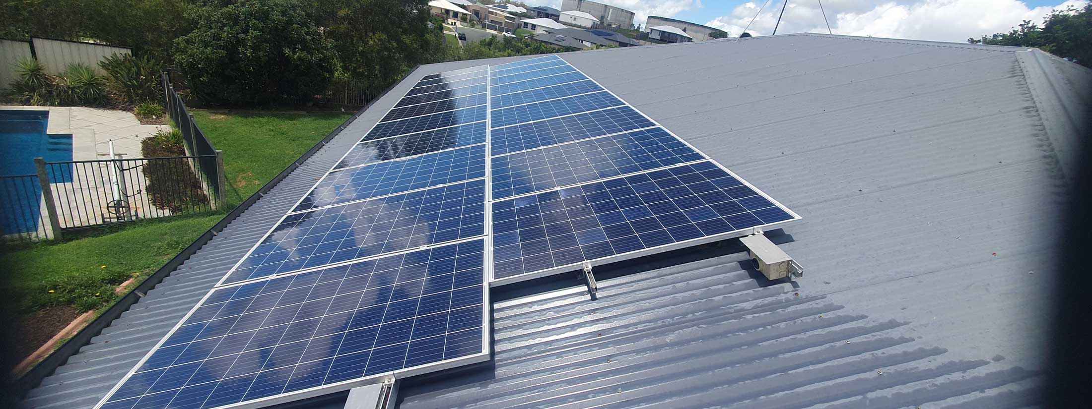 Solar panels mounted on a slanted gray metal roof over a backyard with a pool and fence in view.