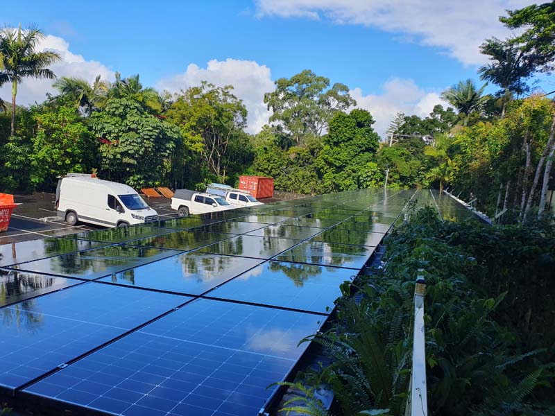 Row of blue solar panels in a parking lot, with white vans and trucks parked beside them and lush green trees in the background under a blue sky.